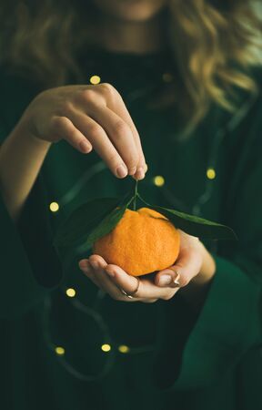 Fresh raw tangerine citrus fruit with leaves in hands of blonde girl wearing green dress with holiday lights bokeh, selective focus, copy space. Winter Christmas of New year holiday mood conceptの写真素材