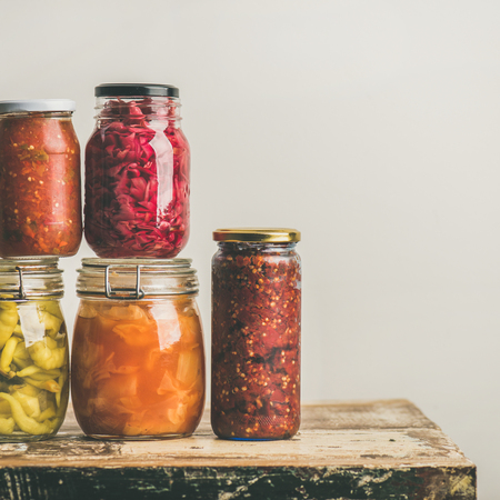 Autumn seasonal pickled or fermented colorful vegetables in glass jars placed in stack over vintage kitchen drawer, white wall background, copy space, square crop. Fall home food preserving or canningの写真素材