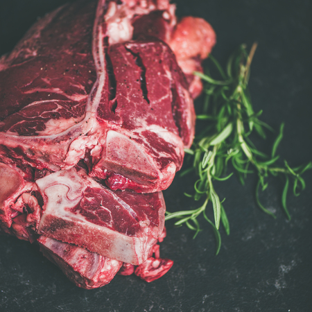 Close-up of fresh raw uncooked beef meat t-bone steaks placed in stack with rosemary over black stone background, selective focus, copy space, square cropの写真素材