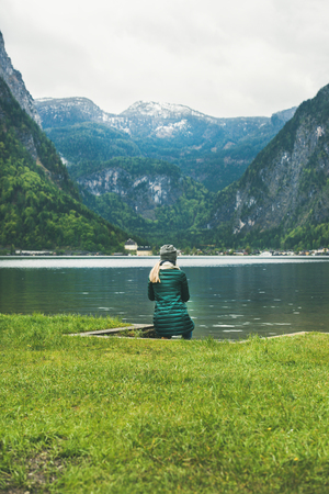 Female traveller in warm clothes from back, sitting at mountain lake coast enjoying the view of the Alps in Austria. Freedom conceptの写真素材
