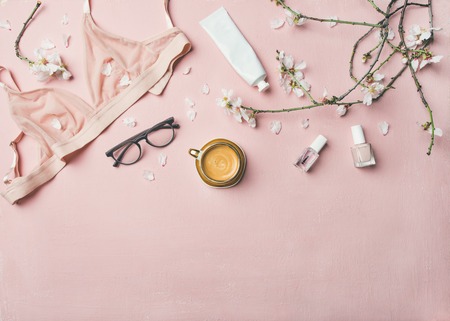 Womans morning rituals concept. Flat-lay of feminine tender powder color lingerie, glasses, cosmetic items, cup of coffee and Spring blossom flowers over pastel pink bed cover, top view, copy space.の写真素材