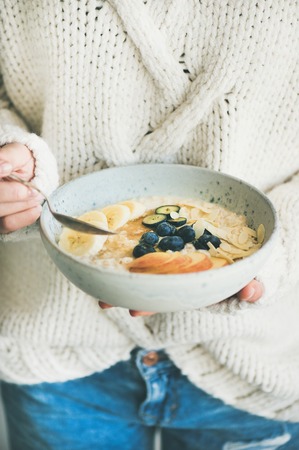 Healthy winter breakfast in bed. Woman in woolen sweater and jeans eating vegan almond milk oatmeal porridge with berries, fruit and almond. Clean eating, vegetarian food conceptの写真素材