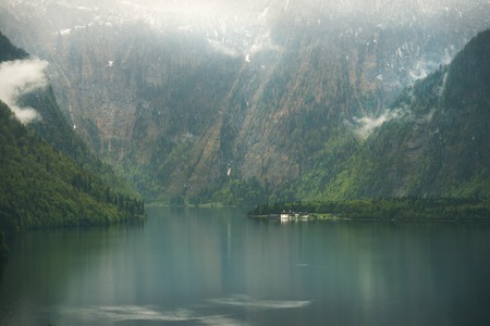 View over misty Lake Konigssee with tiny St Bartholomae church in Berchtesgaden National Park, Bavaria, Germanyの写真素材
