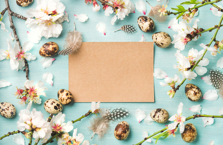 Easter holiday background. Flat-lay of tender Spring almond blossom flowers on branches, feathers, quail eggs and paper in center over blue background, top view, copy space. Greeting card conceptの写真素材