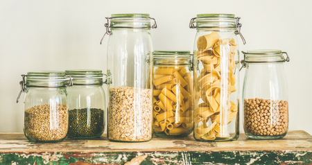 Various uncooked cereals, grains, beans and pasta for healthy cooking in glass jars on rustic table, white background, wide composition. Clean eating, vegetarian, vegan, balanced dieting food conceptの写真素材