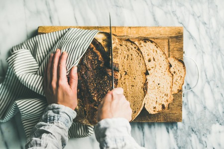 Flat-lay of female hands cutting freshly baked sourdough bread into pieces on rustic wooden chopping board over light grey marble background, top viewの写真素材
