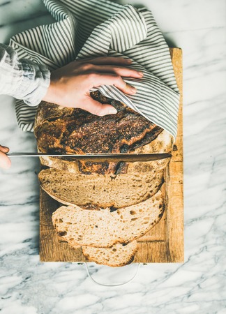 Flat-lay of womans hands cutting freshly baked sourdough bread with knife into pieces on rustic wooden chopping board over light grey marble background, top view, vertical compositionの写真素材