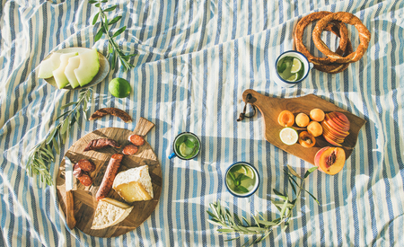 Flat-lay of summer picnic set with fruit, cheese, sausage, bagels and lemonade over striped blanket, top viewの写真素材