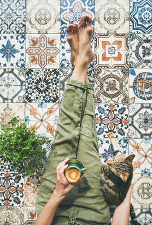 Lazy morning on terrace. Flat-lay of womans legs in cosy linen pants, plant, cat and cup of coffee in hand over colorful moroccan tile floor, top viewの写真素材