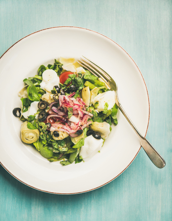 Flat-lay of fresh green summer salad with artichokes, olives, soft cheese and red onion in white plate over blue wooden background, top viewの写真素材