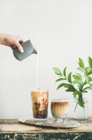 Iced coffee in tall glasses with milk poured over from pitcher by hand, white wall and green plant branches in vase at background, copy space. Summer refreshing beverageの写真素材