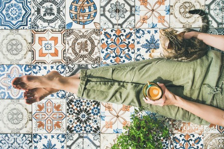 Lazy morning on terrace. Flat-lay of womans legs in cosy linen pants, plant, cat and cup of coffee in hand over colorful moroccan tile floor, top viewの写真素材