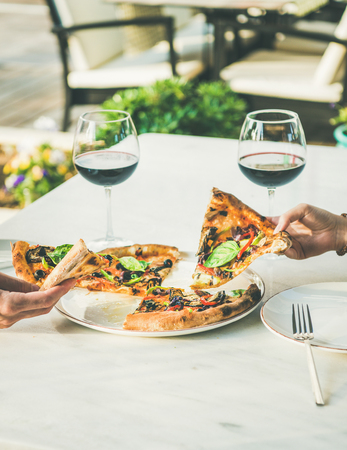 Summer dinner or lunch. People eating freshly baked Italian vegetarian pizza with vegetables and fresh basil and drinking red wine in outdoor restaurant, copy spaceの写真素材