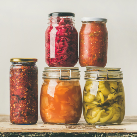 Autumn seasonal pickled or fermented colorful vegetables in glass jars placed in stack over vintage kitchen drawer, white wall background, square crop. Fall home food preserving or canningの写真素材