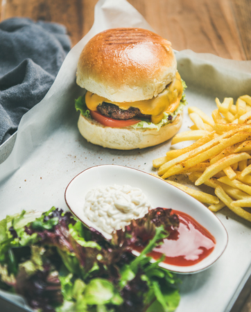 Classic burger dinner. Beef meat homemade burger with French fries, salad and sauces on white tray over rustic wooden table background, selective focusの写真素材