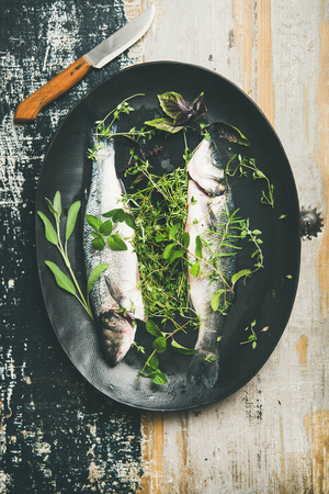 Cooking fish dinner. Flat-lay of raw uncooked sea bass fish with fresh herbs on dark plate over rustic wooden painted background, top view. Helathy, clean eating, dieting food conceptの写真素材