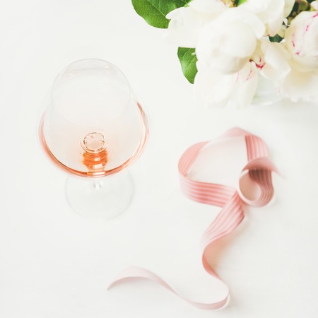Rose wine in glass, pink decorative ribbon, peony flowers over white table background, copy space, square crop. Summer celebration, wedding greeting card, invitation conceptの写真素材