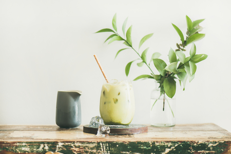 Iced matcha latte drink in glass with coconut milk pouring from pitcher by womans hand, white wall and plant branches at background, horizontal composition. Summer refreshing beverage cold drinkの写真素材