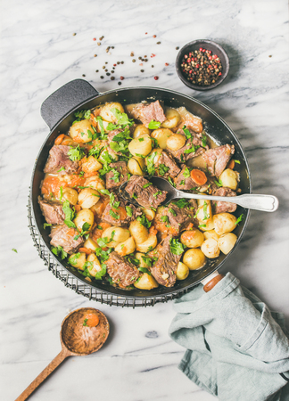 Meat dinner. Flat-lay of braised beef meat with potato and carrot with fresh parsley in cooking pan over white marble table background, top view. Comfort winter foodの写真素材