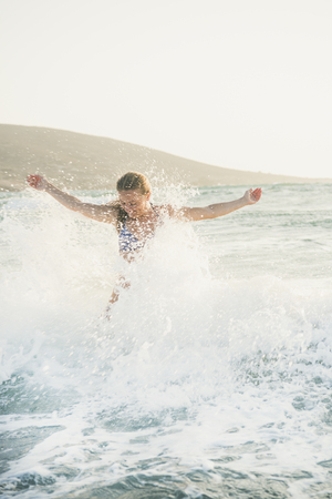 Young beautiful blond woman tourist in swimsuit standing and enjoying splash of wavy waters of Mediterranean sea in Prasonisi cape, Rhodes, Greece on summer dayの写真素材