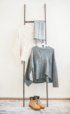 Fall or winter warm knitwear on hanger, white wall background. Fashionable female winter clothing. Woolen sweaters, grey scarf and winter bootsの写真素材