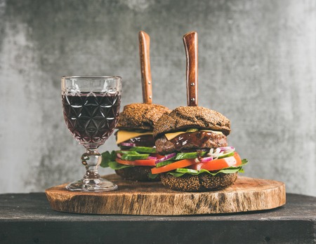 Beef meat cheeseburgers with barbeque sauce and glass of red wine on rustic wooden board, grey concrete wall at background. Comfort food, burger bistro conceptの写真素材