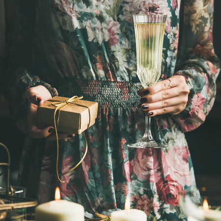 Woman in festive dress holding glass of champaigne and gift box in hands at holiday decorated table during Christmas or New Year celebration party, selective focus, square cropの写真素材