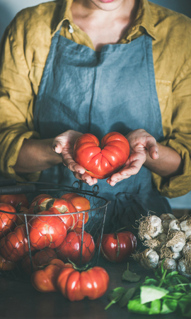 Woman in linen apron holding ripe heirloom tomato for cooking tomato sauce, canned tomatoes or pasta with basil and garlic at kitchen counter. Healthy cooking, slow food or comfort foodの写真素材