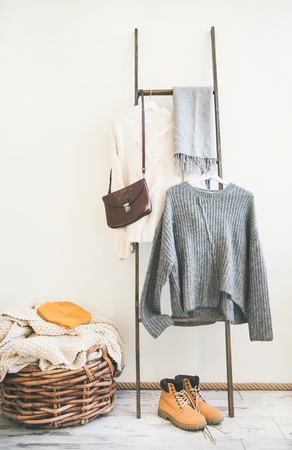Fall or winter warm knitwear on hanger, white wall background. Fashionable female winter clothing. Woolen sweaters, scarf, boots, shoulder bag and knitted cap, copy spaceの写真素材
