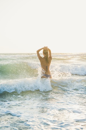 Young blond woman tourist in swimsuit standing backwards and enjoying wavy waters of Mediterranean sea in Prasonisi cape, Rhodes, Greece on summer day at sunsetの写真素材