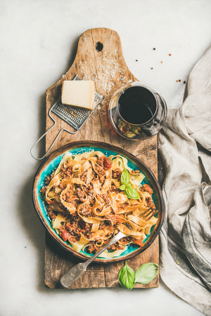Italian traditional pasta dinner. Flat-lay of Tagliatelle bolognese with minced meat, tomato sauce and parmesan cheese and glass of red wine over rustic wooden board, white table background, top viewの写真素材