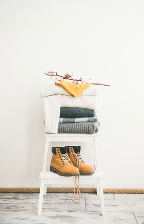 Heap of knitted warm blankets, scarves and sweaters, cap and boots for winter or fall cold weather and tree branch on white stool near white wall, copy space. Cosy home winter atmospereの写真素材