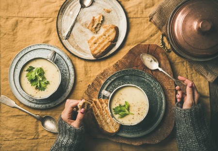Autumn or Winter home dinner. Flat-lay of Fall warming celery cream soup in cups and female hands in sweater over linen tablecloth, top view. Comfort food, vegetarian, healthy and slow food conceptの写真素材