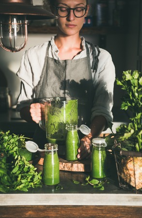 Making green detox take-away smoothie. Woman in linen apron and glasses pouring green smoothie drink from blender to bottle surrounded with vegetables and greens. Healthy, weight loss food conceptの写真素材