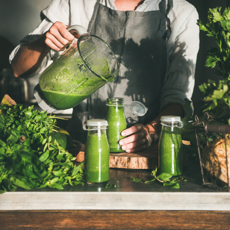 Making green detox take-away smoothie. Woman in linen apron pouring green smoothie drink from blender to bottle surrounded with vegetables and greens, square crop. Healthy, weight loss food conceptの写真素材