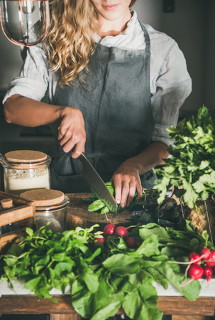 Fall seasonal vegetarian, vegan dinner cooking. Woman in linen apron cutting green vegetable ingredients on concrete kitchen counter. Slow food, comfort food, healthy diet, clean eatingの写真素材