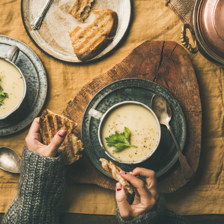 Autumn, Winter home dinner. Flat-lay of Fall warming celery cream soup and female hands with bread over linen tablecloth, top view, square crop. Comfort food, vegetarian, healthy and slow food conceptの写真素材