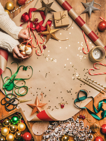 Preparing for New Year. Flat-lay of hot chocolate in mug in womans hands, candy canes, decorations, Christmas ribbons, wreath, glittering balls over gift paper background, top view, copy spaceの写真素材