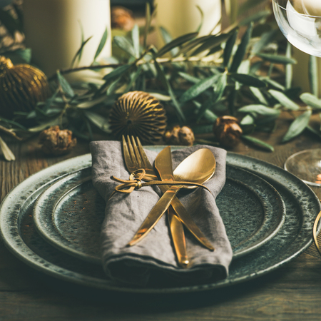 Christmas or New Years celebration party table setting. Plates, golden cutlery, glasses, gift box, festive branch decoration, candles, gliterring toys over rustic wooden table background, square cropの写真素材