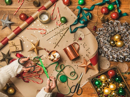 Preparing for New Year, Chrismtas. Flat-lay of hot chocolate with marshmallows, woman's hands cutting Christmas ribbons, door wreath, glittering balls, candy canes over gift paper, top viewの写真素材