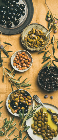 Flat-lay of various kinds of Mediterranean pickled olives in plates and bowls and olive tree branches on yellow linenの写真素材