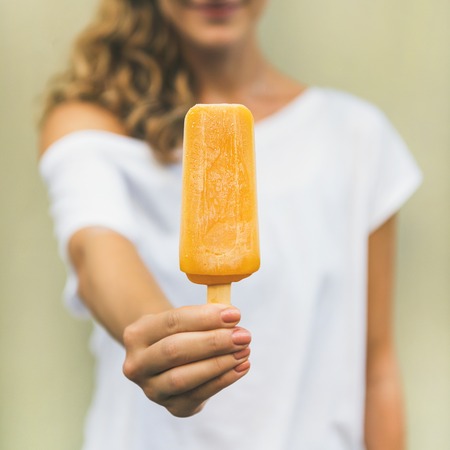 Healthy vegan orange mango citrus ice cream popsicle in hand of young woman with yellow wallの写真素材