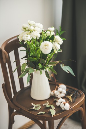 Spring white buttercup flowers in enamel jug on wooden vintage chair with white wall and black curtain