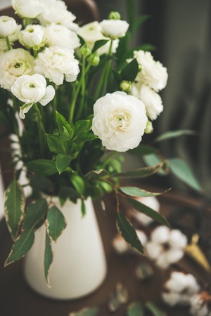 Spring white buttercup flowers in white enamel jug on wooden vintage chair with curtain behind, close-up. Wedding bouquet, flower shop, Womens Day holiday or spring mood conceptの写真素材