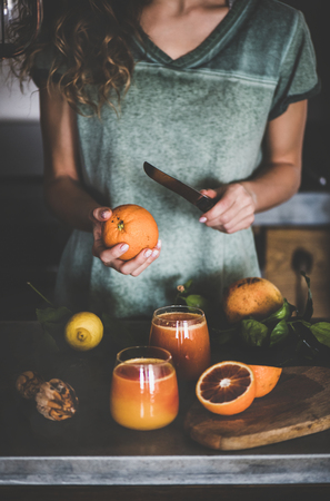 Young female making freshly squeezed blood orange juice or smoothie near concrete kitchen counter. Healthy lifestyle, vegan, vegetarian, alkaline diet conceptの写真素材