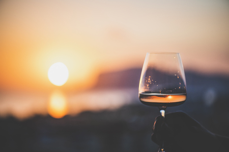 Man's hand holding glass of rose wine and with sea and beautiful sunset at background, close-up, horizontal composition. Summer evening relaxed mood concept