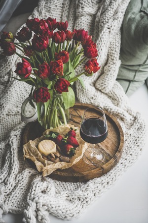 Wine snack set with flowers. Glass of red wine, cheese, roasted almonds, strawberries and bouquet of fresh Spring red tulips on wooden tray over white knitted blanket. Romantic or relaxed mood conceptの写真素材