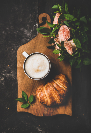 Coffee break. Flat-lay of cup of hot cappuccino with croissant and bouquet of pink flowers over black shabby background, top view, vertical composition. Spring moodの写真素材