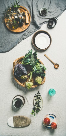 Flat-lay of spring table setting with fresh vegetables, fruits, olive oil and tableware over light grey linen tablecloth, top view, vertical compositionの写真素材