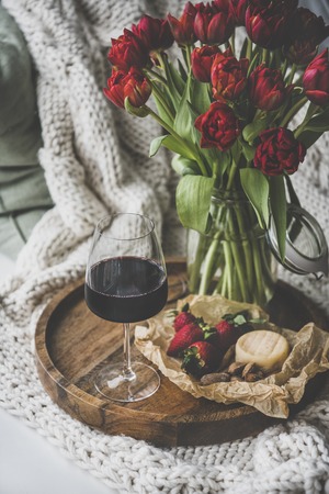 Wine snack set with flowers. Glass of red wine, cheese, roasted almonds, strawberries and bouquet of Spring red tulips on wooden tray over white knitted blanket. Romantic mood conceptの写真素材